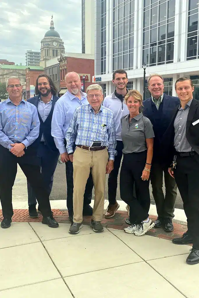 An outdoor photo shows eight people—six men and two women—standing side-by-side on a sidewalk in an urban setting. They are all casually dressed, though some are in suits/sport coats and others are in more relaxed attire. A tall, older man in a blue and white plaid shirt and tan pants is centered in the group. In the background, there is a modern glass-front building and, further in the distance, an older building with a large dome and clock tower. The group is from Bradley Company in Fort Wayne, Grand Rapids, and Indianapolis.
